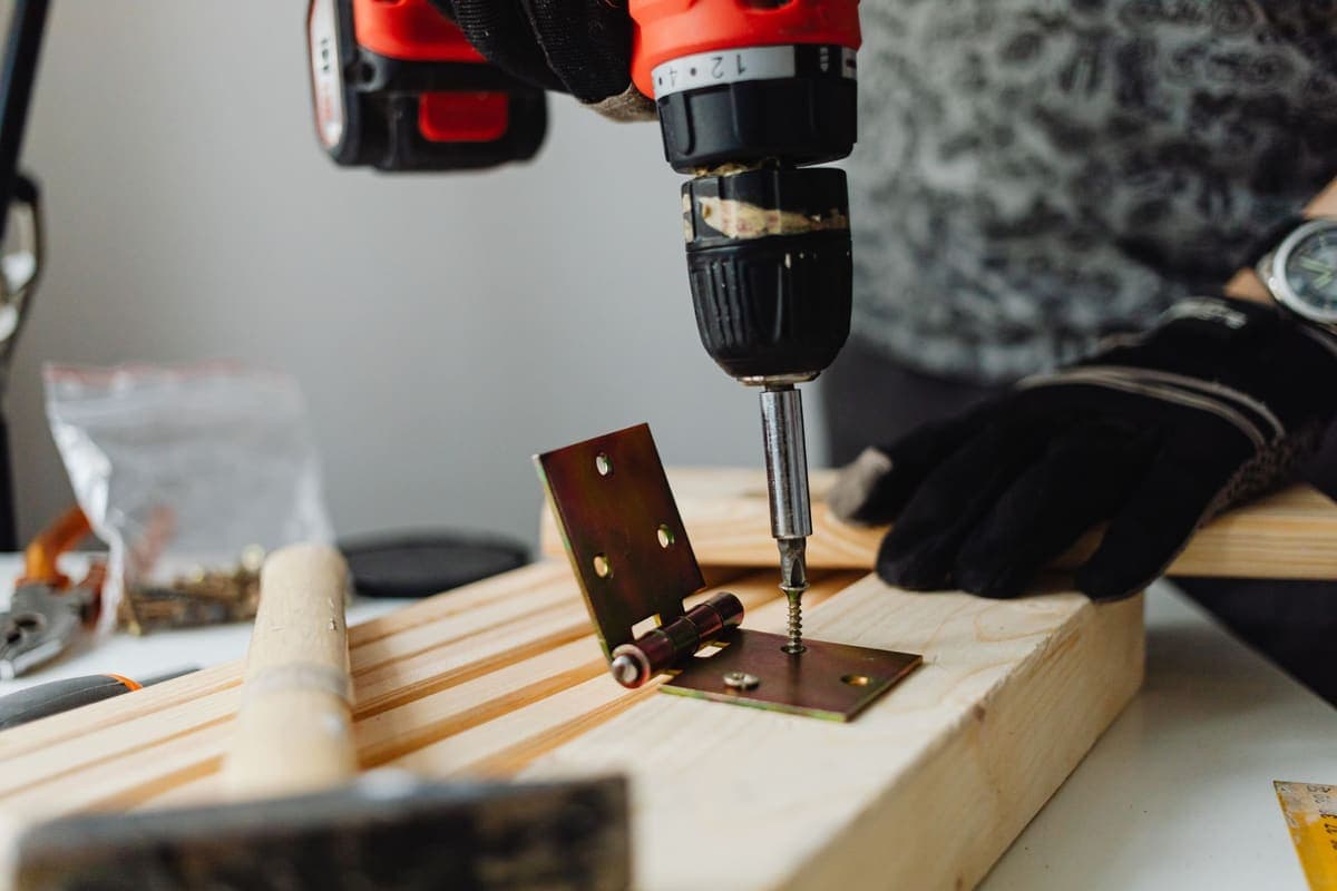 A cordless drill driving a screw through a hinge plate into wood